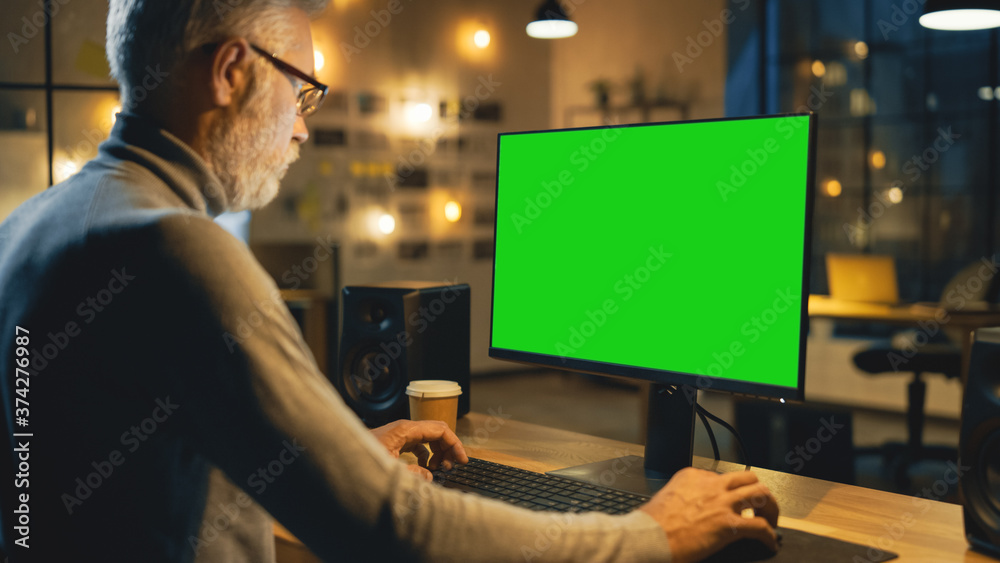 富有创意的中年设计师坐在办公桌前使用带有绿色实体屏幕的台式电脑。I