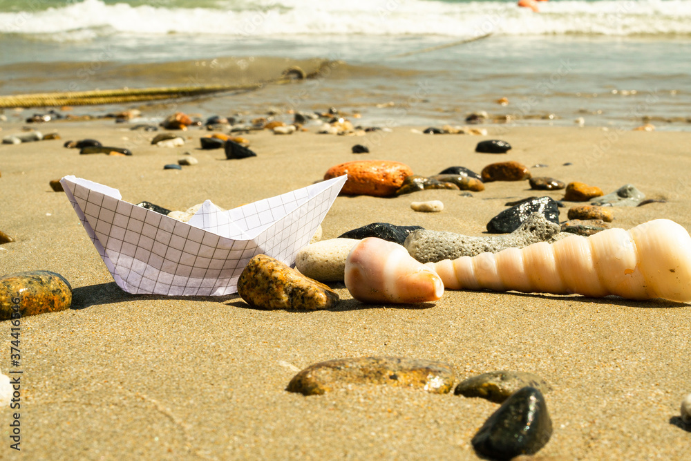在海浪的背景下，沙滩上的纸船在贝壳和石头之间。复制空间。
