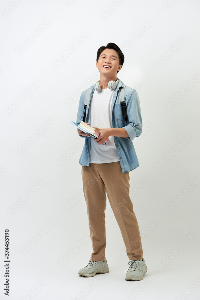 快乐的年轻亚洲学生，带着书、耳机、背包，随意地站在白色的背上