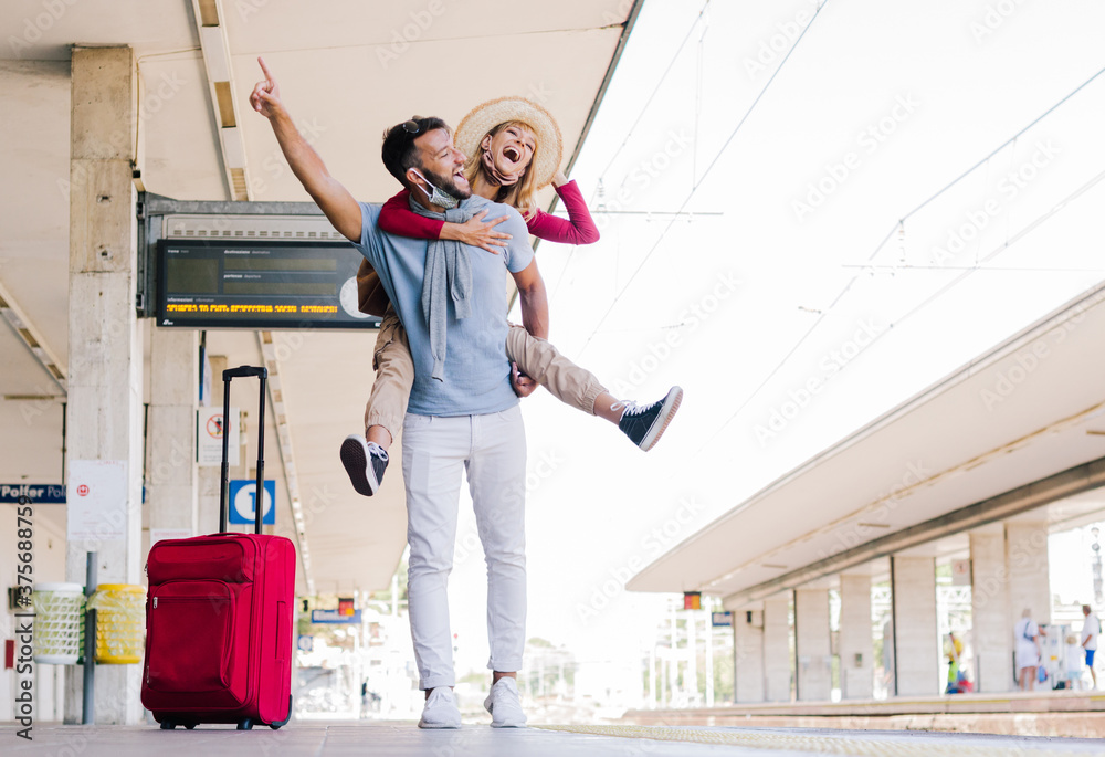 Happy couple in love wearing face mask having fun at train station. New normal travel concept.