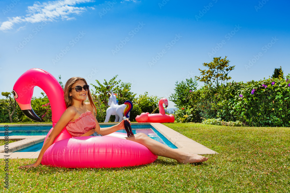 漂亮的女孩，手里拿着苏打水，戴着太阳镜，躺在游泳池附近的粉红色火烈鸟上