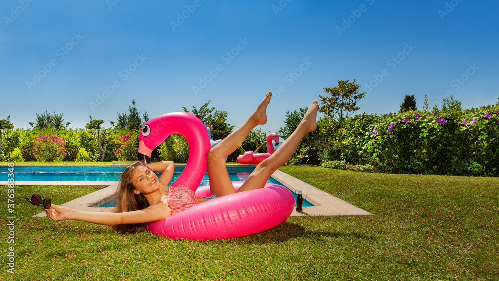 美丽的女孩在一个有趣的动作中摔倒，戴着太阳镜躺在游泳池附近的玫瑰火烈鸟上