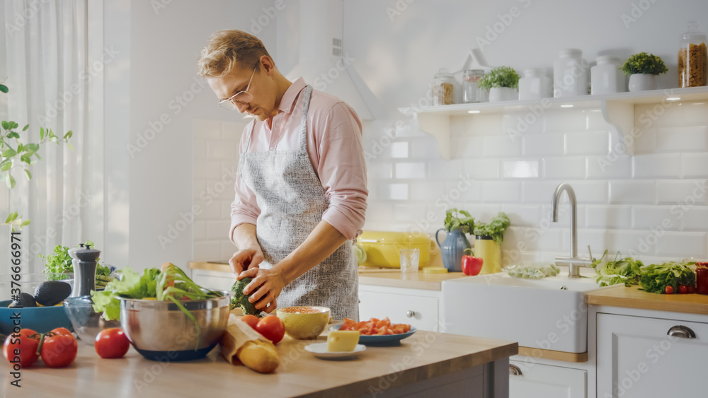 穿着粉色衬衫和围裙的英俊男子正在现代阳光厨房制作健康的有机沙拉餐