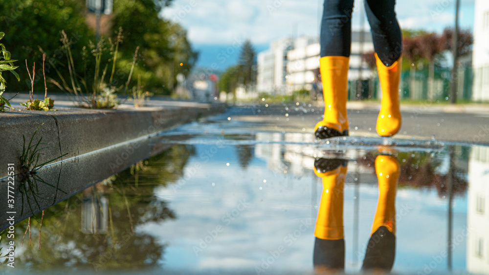 低角度：一个穿着黄色雨靴的女人跑进了一个玻璃般的大水坑。