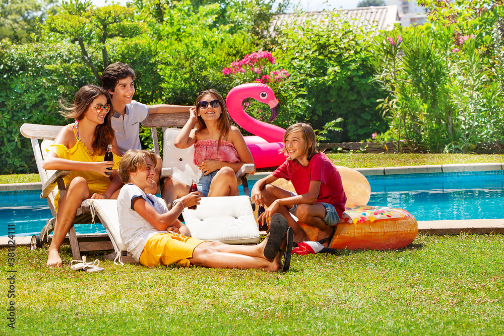 Group of happy teenagers kids drinking soda chatting sit by the pool near deck chair