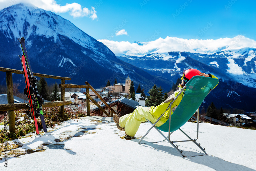 Young woman rest in the deck chair near pair of ski observing mountains and Alpine peaks