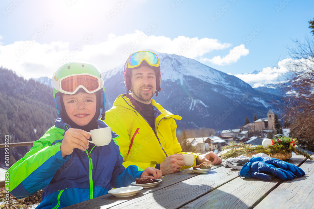 Little child boy in ski outfit drink tea with father in cafe over mountain view panorama