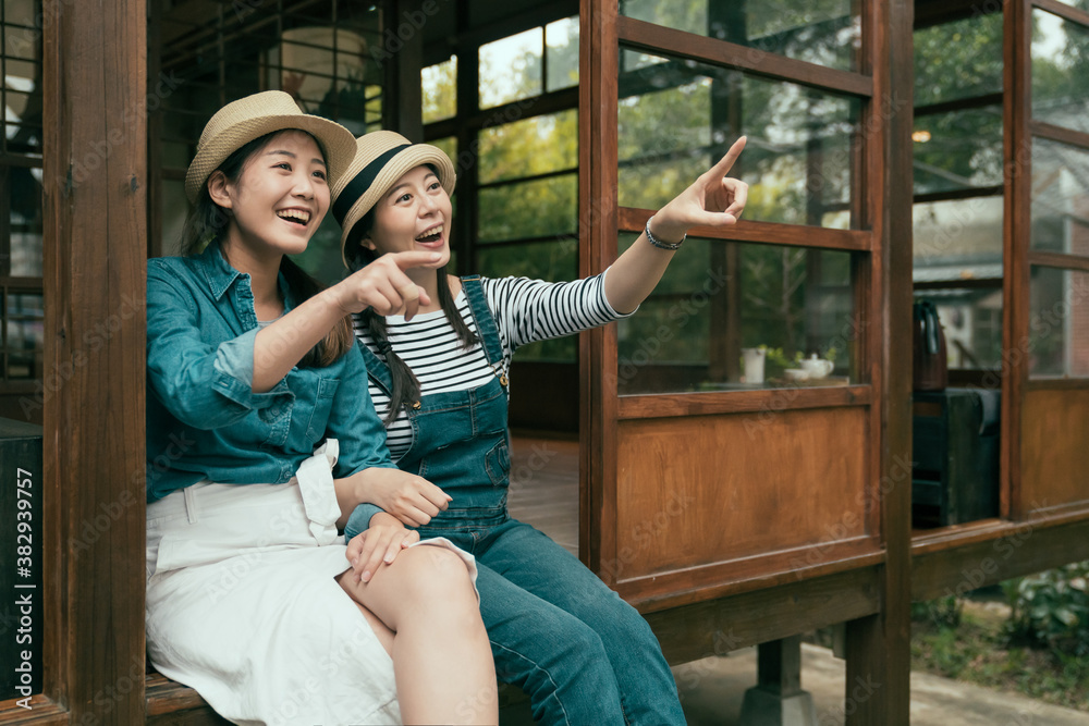 two cheerful young asian chinese girl travelers pointing finger away while relax sitting on wooden f