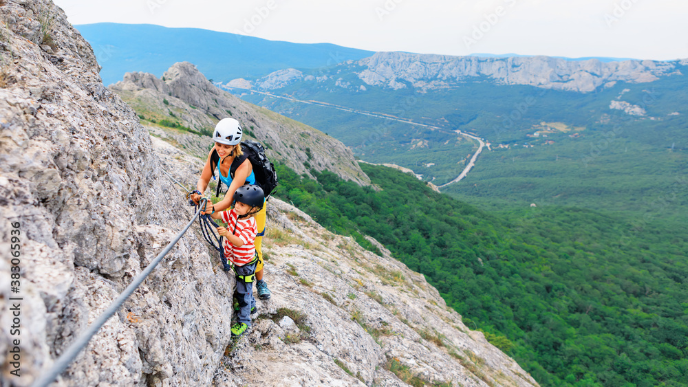 Young mother, child in safety equipment climb to mount top by via ferrata beginner route. Family tra