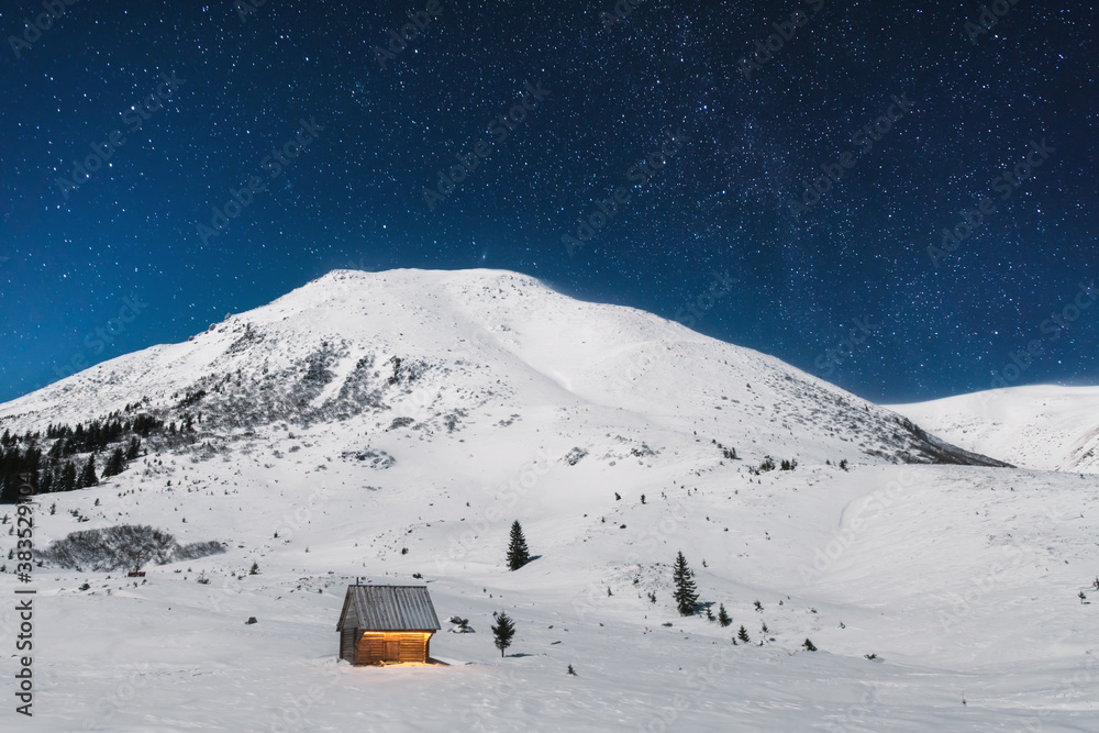 Fantastic winter landscape with wooden house in snowy mountains. Starry sky with Milky Way and snow 