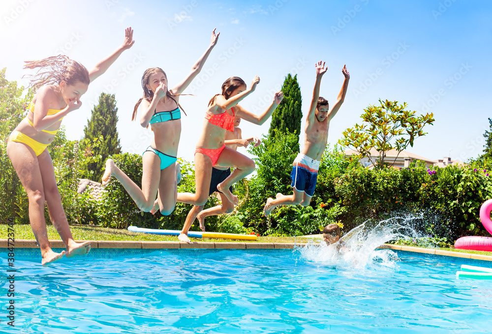 Happy kids in a group of friends jump into water of swimming pool together splashing down and liftin