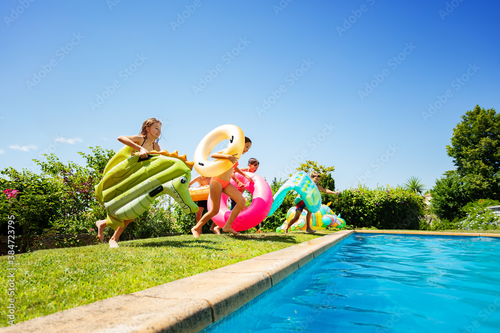 Group of many young kids run and jump into the swimming pool holding inflatable buoy toys diving in 