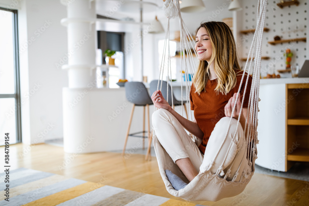 Young woman chilling at home in comfortable hanging chair in front of big window.