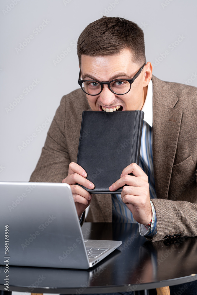 Guy bites closed notebook. Businessman with busy face and glasses isolated on light gray background.