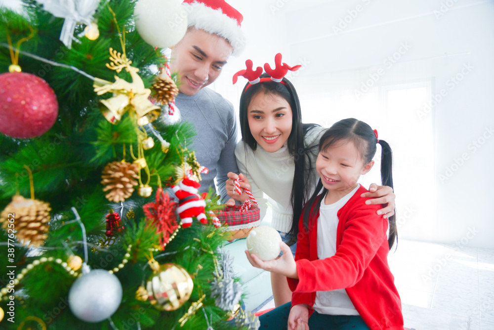 Happy asian parents and their little daughter decorating christmas tree in their house.