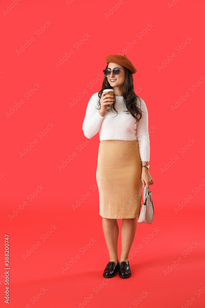 Stylish young woman in autumn clothes and with cup of coffee on color background