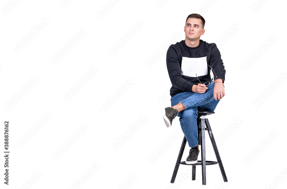 Smiling guy sits on high chair with leg on knee. White background and casual clothes. Copy space.