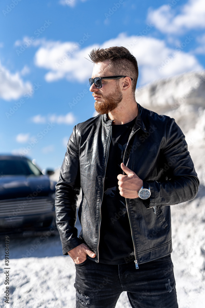 黑色汽车旁站着一位戴着时髦墨镜、穿着皮夹克的时尚胡须男子的肖像。Fas