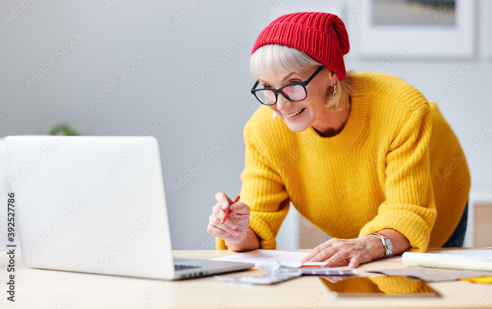 戴着红帽子的开朗的老年女性创意设计师拿着笔记本电脑在工作场所微笑，ske