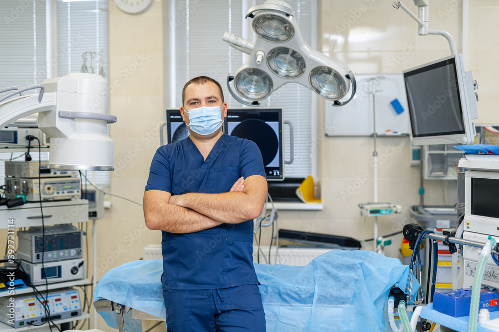 一位戴着口罩、双臂交叉站在手术室里的男外科医生的肖像。专业而自信