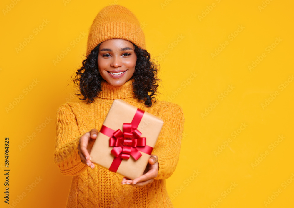 cheerful smiled  ethnic woman  in yellow sweater and hat  holds a Christmas gift on colorful   backg