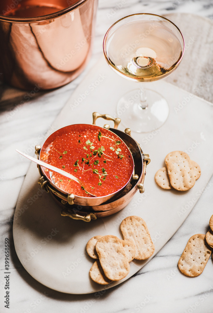Red caviar in copper metal dishes with salty crackers and glass of champaigne over white marble tabl