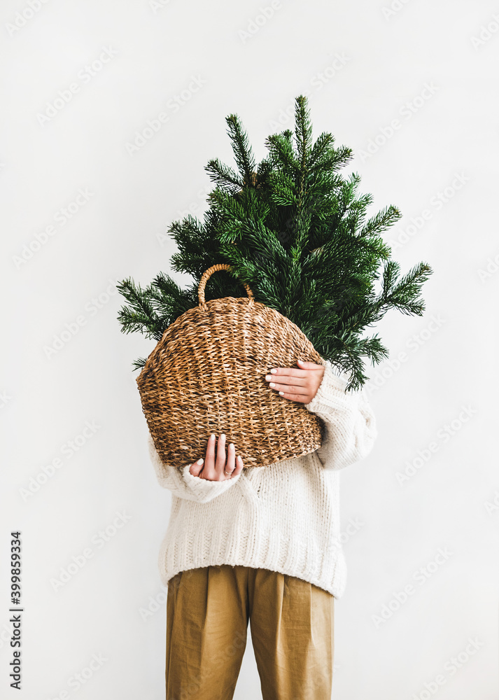 Female figure in white woolen winter sweater and khaki pants standing with wicker bag with evergreen