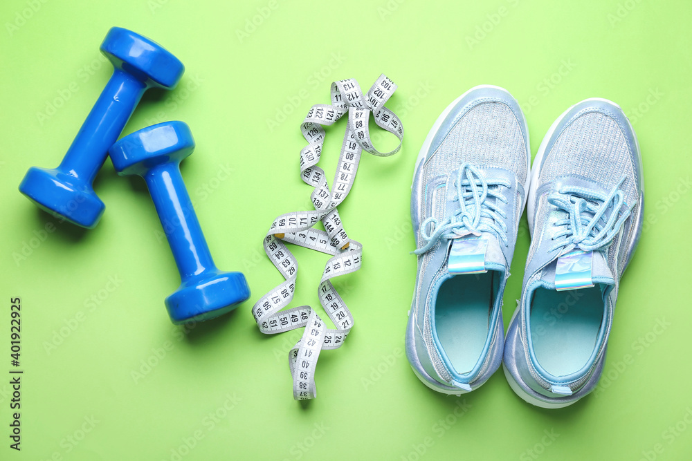 Sportive shoes, dumbbells and measuring tape on color background