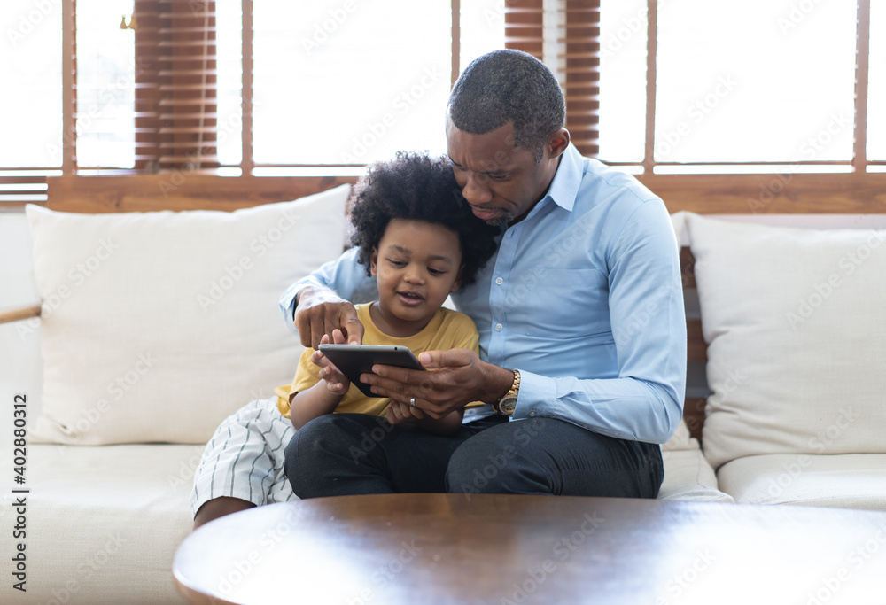快乐的非裔美国人父子坐在家里的沙发上使用平板电脑的肖像。
