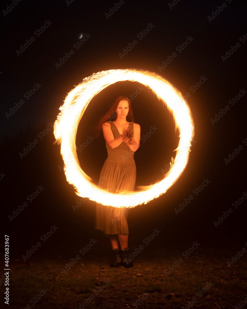 Graceful elegant red haired female fireshow performer making fire dance with two metal fans lit with