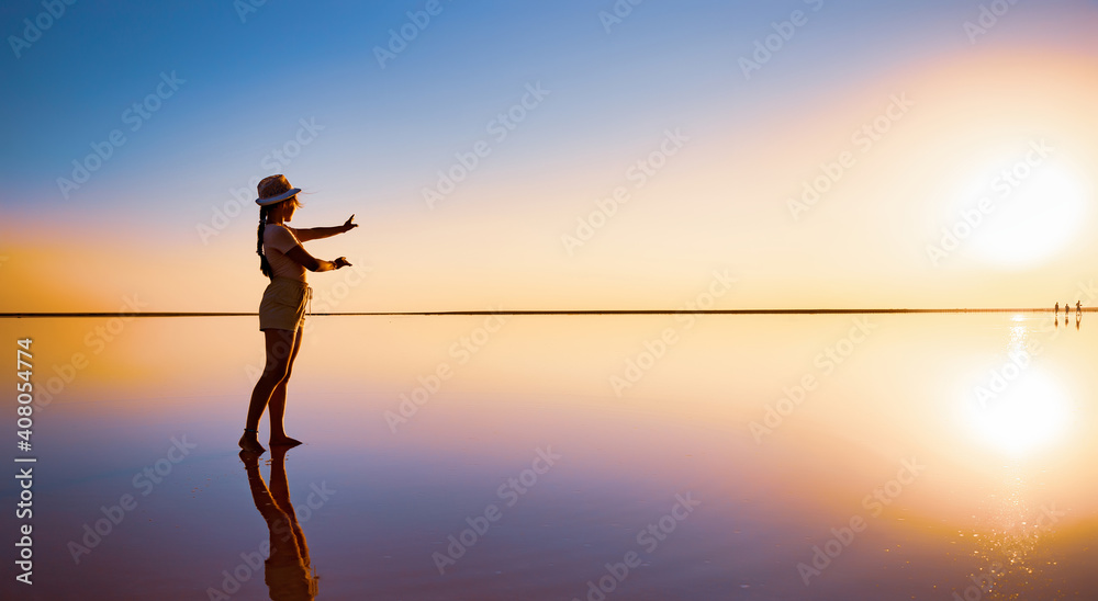 可爱的快乐的年轻女孩沿着镜子般的粉色盐湖边散步，摆出神奇的姿势，享受阳光