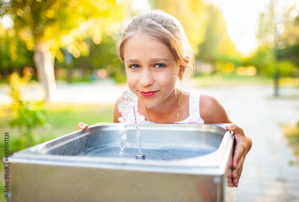 Cheerful wonderful girl drinks cool fresh water from a small fountain in a summer warm sunny park on