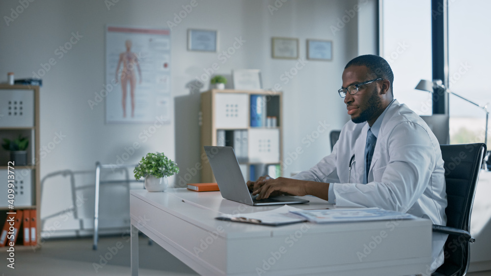 冷静的戴眼镜的非洲裔美国家庭医生正在健康中心的笔记本电脑上工作