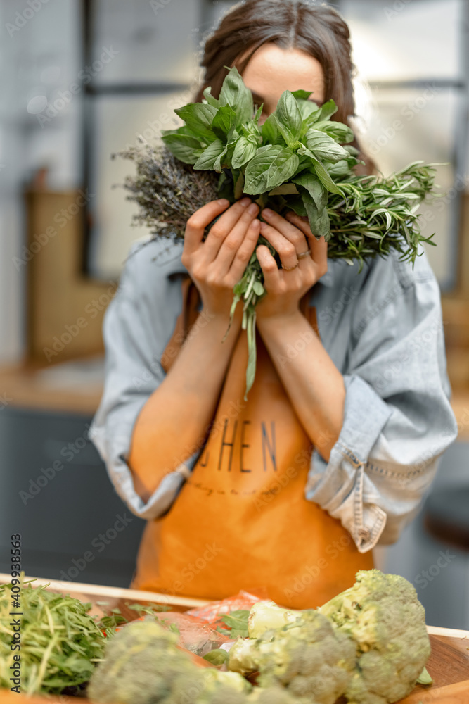 Portrait of cheerful woman in apron with fresh spicy herbs basil, rosemary, thyme on the kitchen. He