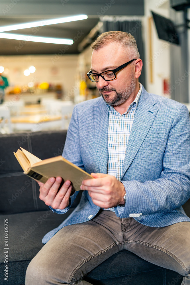 穿着蓝色夹克的男子在咖啡馆看书。戴着时尚眼镜的资深男性坐在沙发上。特写。