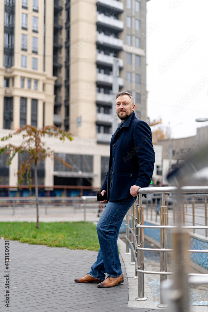 Full body portrait of a fashion man standing outdoors near iron railing. Elegant coat and jeans. Mod