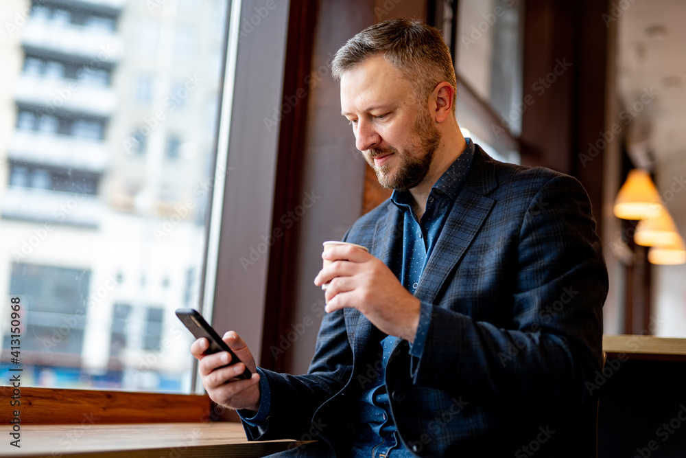 Young businessman in jacket surfs interenet on a phone while resting in a cafe with a cup of coffee.