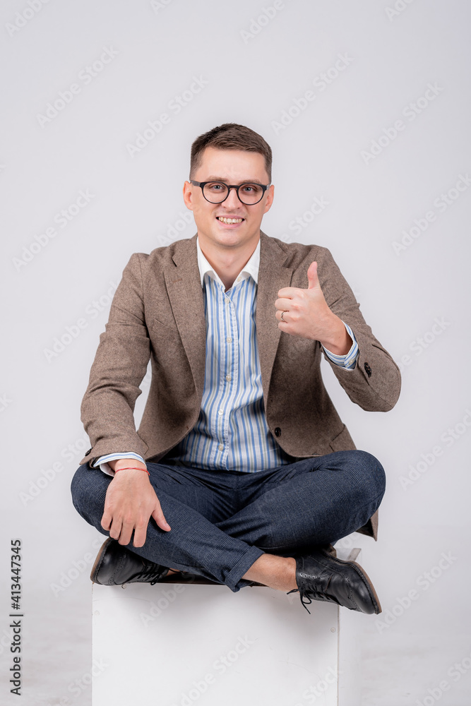 Formal young man sitting cross legged showing thumbs up with one hand and smiling to the camera. Clo