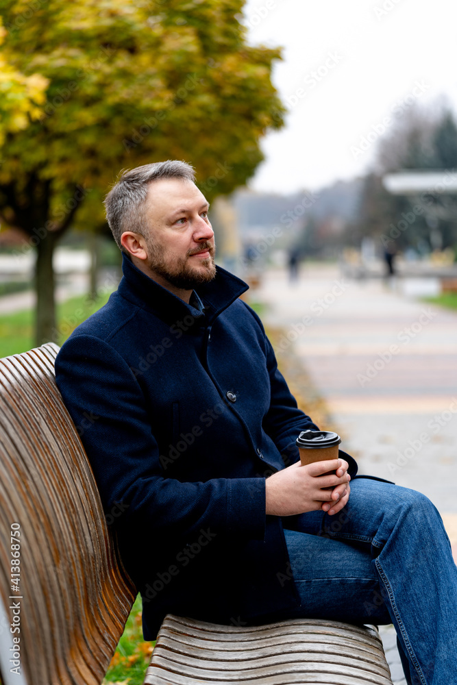 穿着外套的英俊男子手里拿着咖啡杯。秋天，一个男人坐在公园的长椅上。特写。