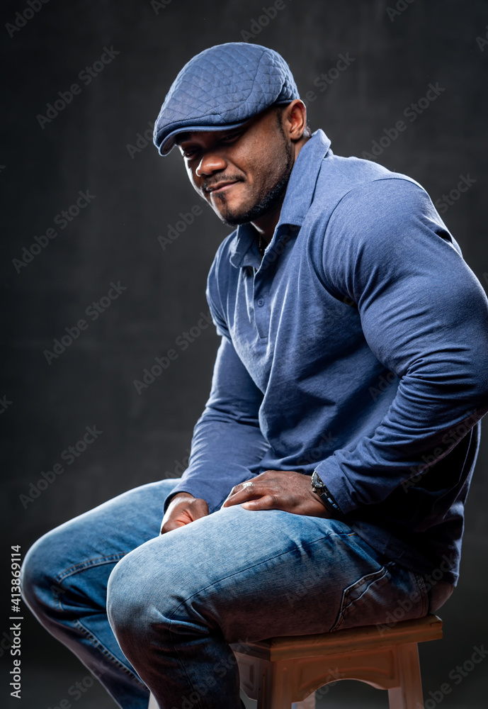 Strong african american in total denim clothes sits on chair and poses to the camera on black backgr