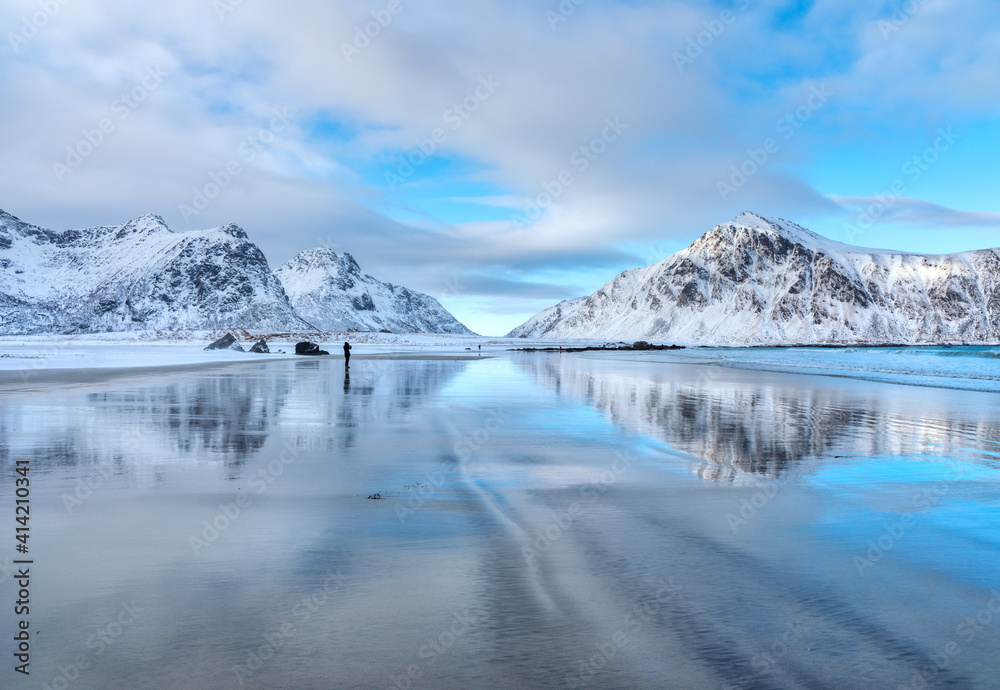 Snowy mountains and blue sky with clouds reflected in water in winter. Arctic sandy beach in Lofoten