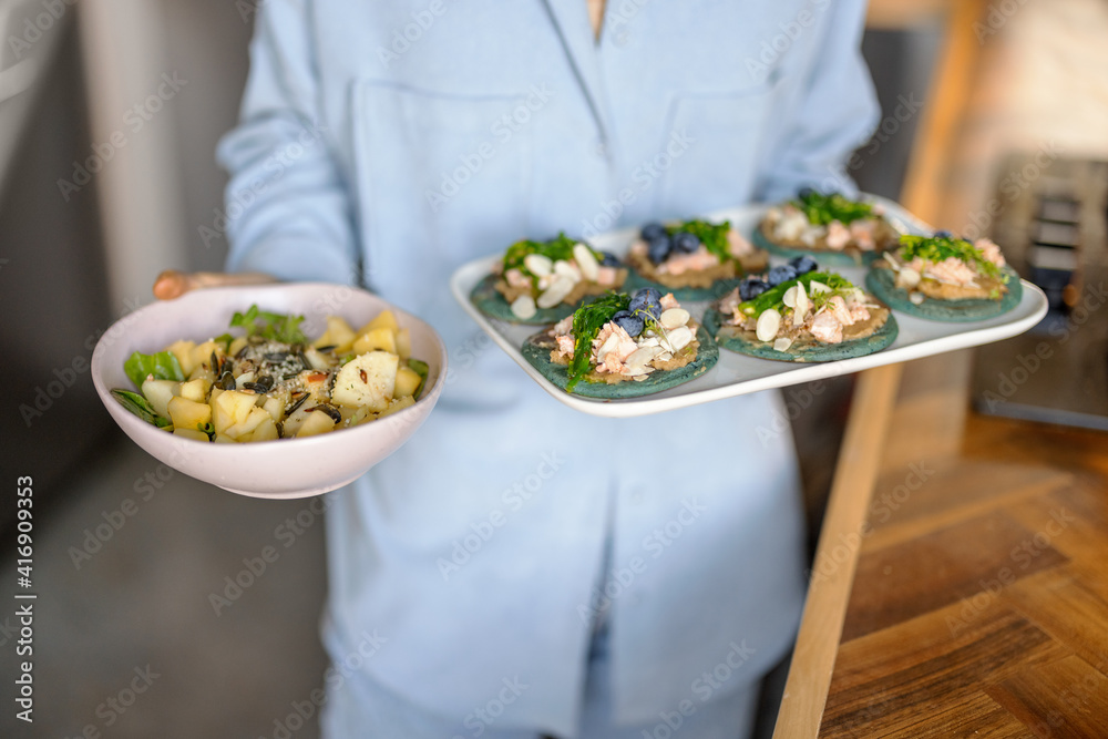 穿着蓝色衬衫的女人在盘子里放着一套开胃菜，里面有海藻、奶酪、蓝莓和鹰嘴豆泥。
