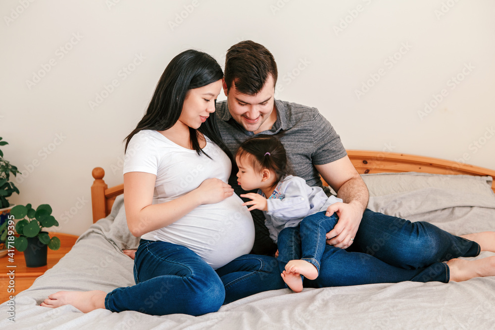 家庭亚裔中国孕妇和高加索男子带着蹒跚学步的女孩坐在家里的床上。Mot