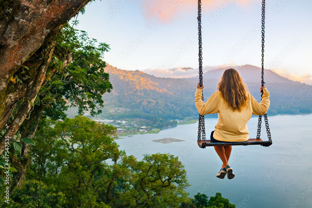 Summer vacation. Young woman sit on tree rope swing on high cliff above tropical lake. Happy girl lo