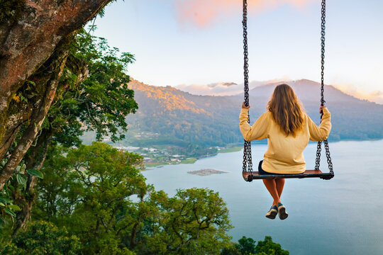 Summer vacation. Young woman sit on tree rope swing on high cliff above tropical lake. Happy girl lo