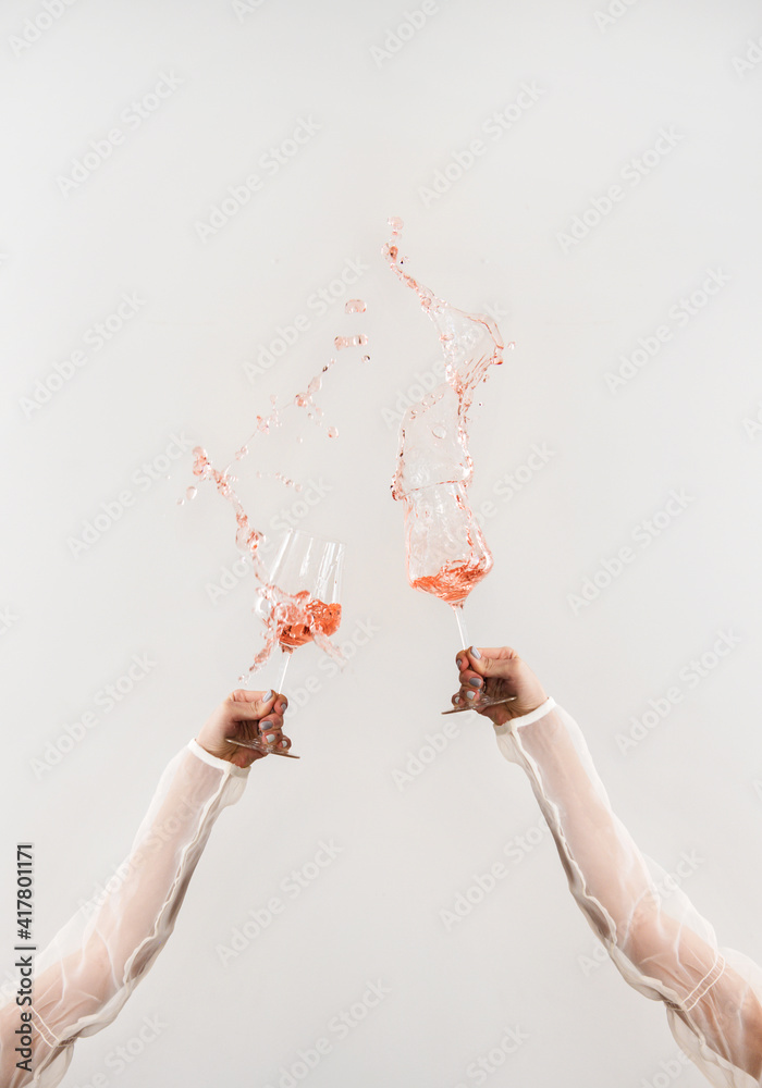 Two hands in white blouse clinking glasses of rose wine making splashes over white wall background. 