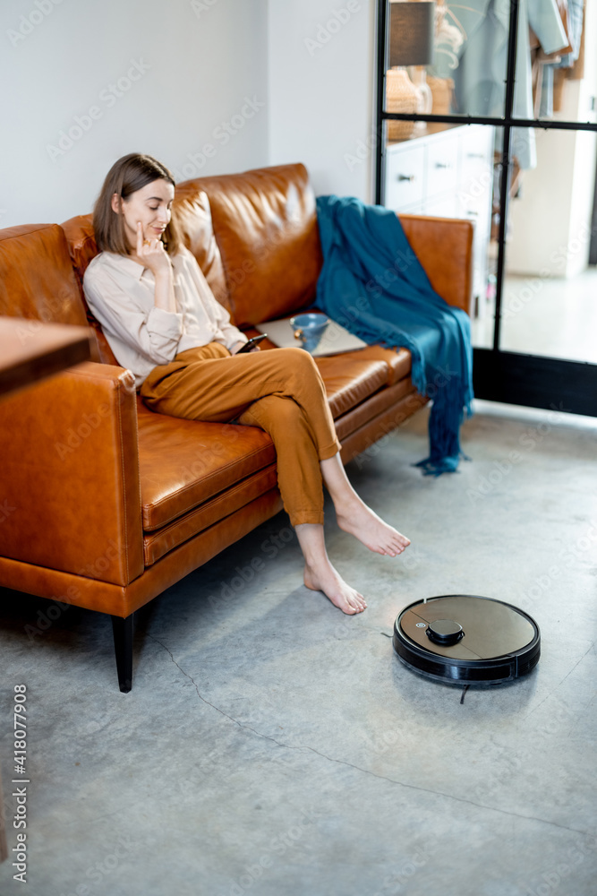 黑色机器人吸尘器正在清洁地板，而漂亮的女人坐在家里的棕色沙发上。L