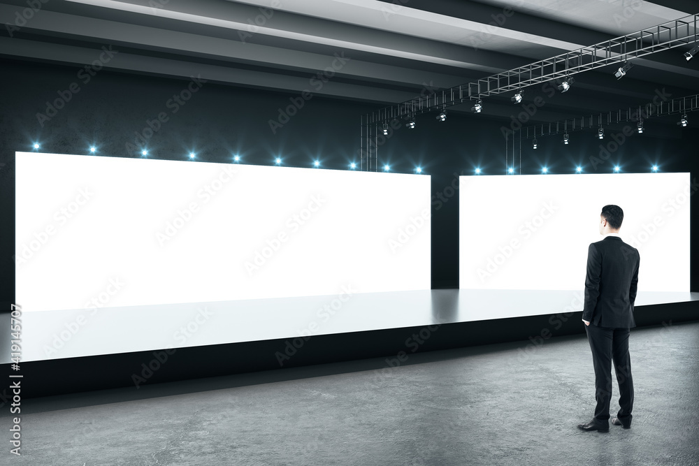 商人在空荡荡的大厅里看着顶部有灯的空白白色大屏幕，屏幕上有黑色光泽的podiu