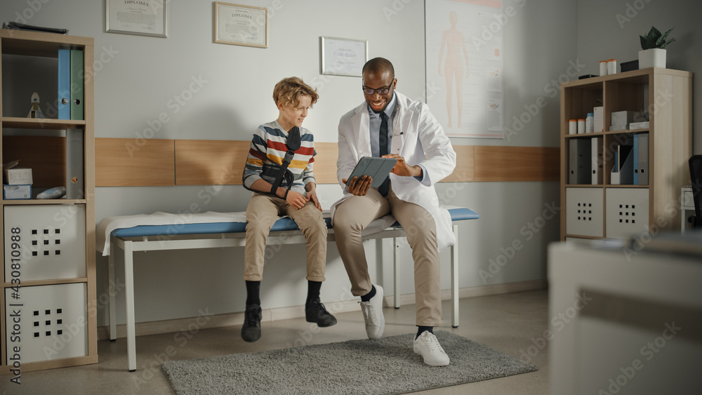 友好的非裔美国家庭医生与一个戴着臂膀的男孩交谈并展示测试结果