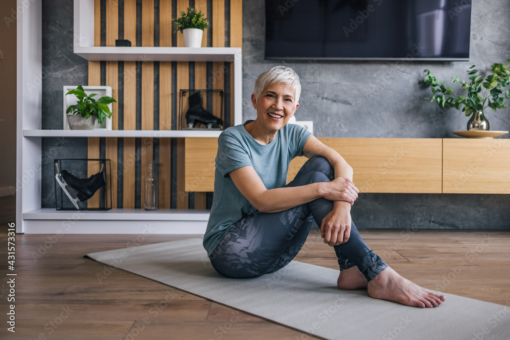 穿着运动服的健康老年女性坐在家里的垫子上，对着镜头微笑。